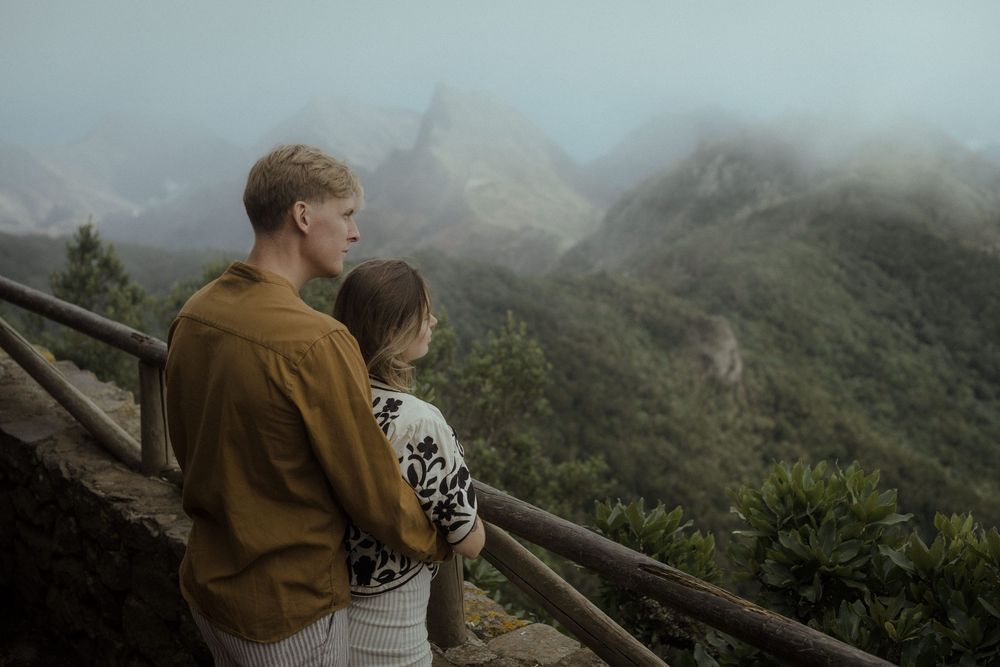 Two people stand on a mountain overlook gazing at misty peaks and forested slopes in moody weather.
