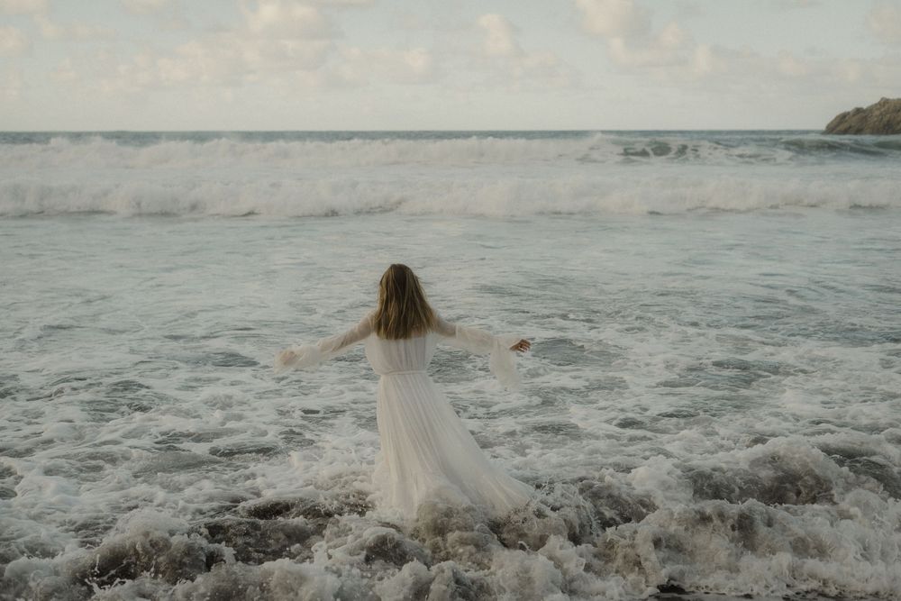 A person in a flowing dress stands at the ocean's edge as waves crash against the rocky shore at sunset.