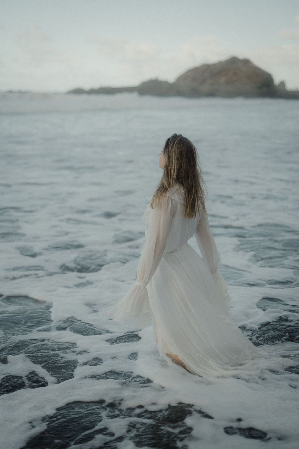 A figure in a flowing white dress stands on a misty beach with rocky cliffs in the background.