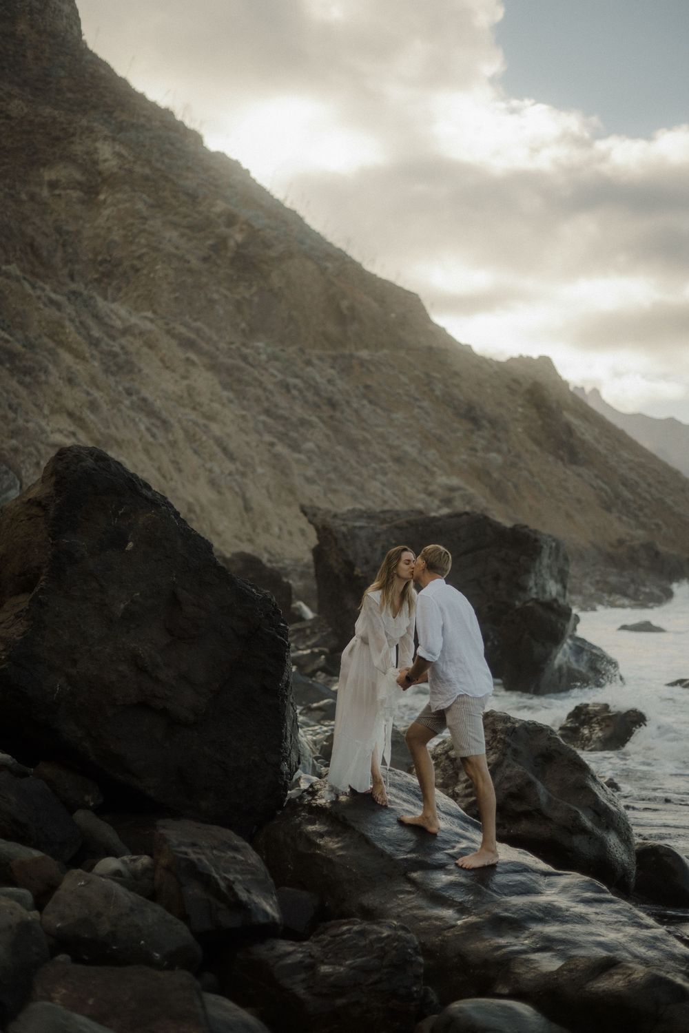 A couple in white outfits share a romantic moment on dark coastal rocks at sunset.