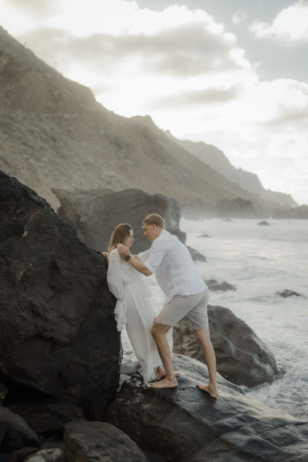A couple in white attire shares an intimate moment on rocky coastal cliffs with dramatic ocean waves in the background.