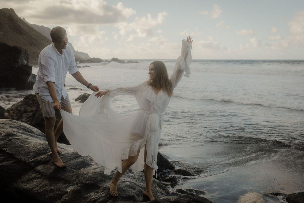 A couple walks along a rocky beach at sunset with waves crashing against the shore.