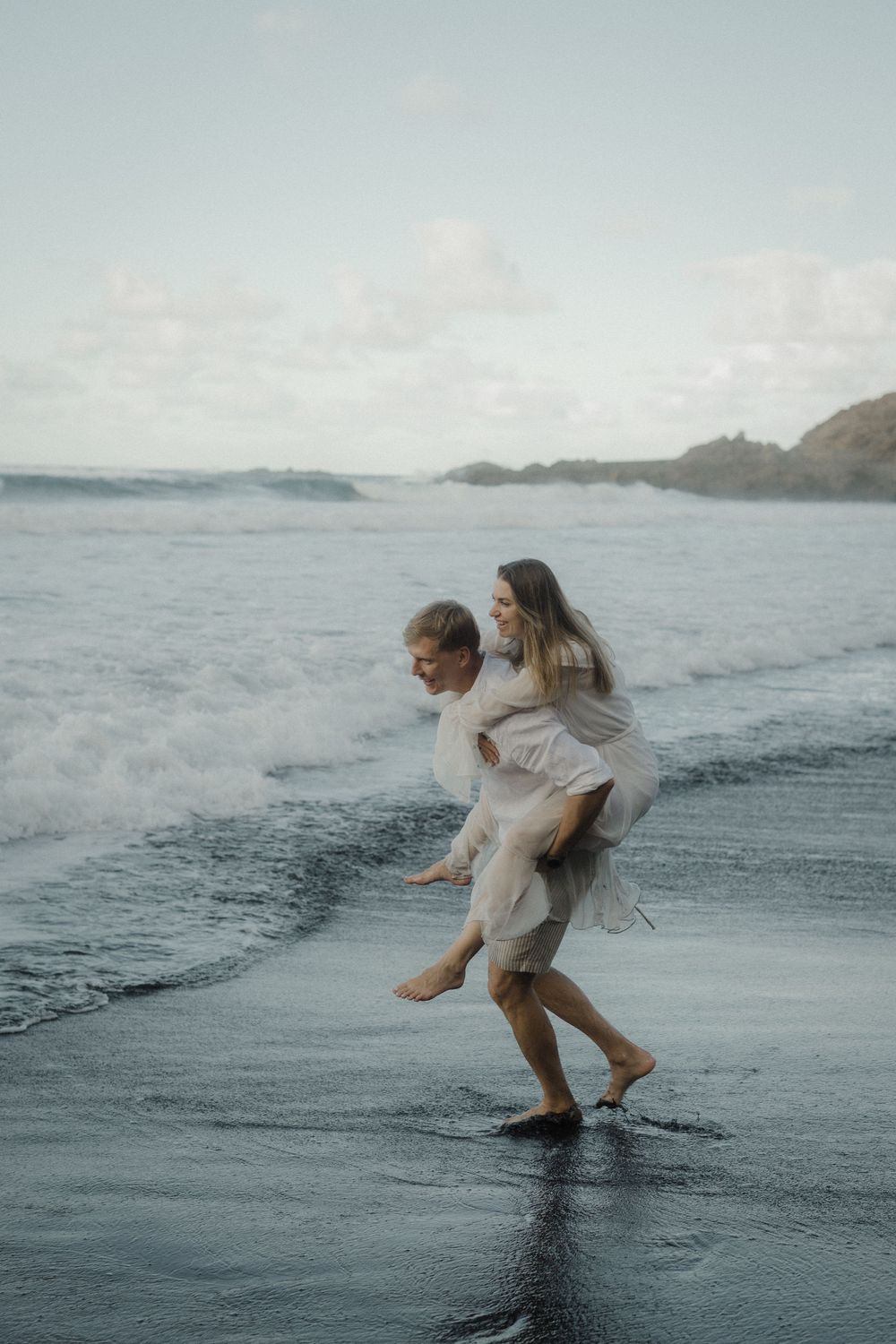 A playful couple runs along a misty beach shore with crashing waves and coastal cliffs in the distance.