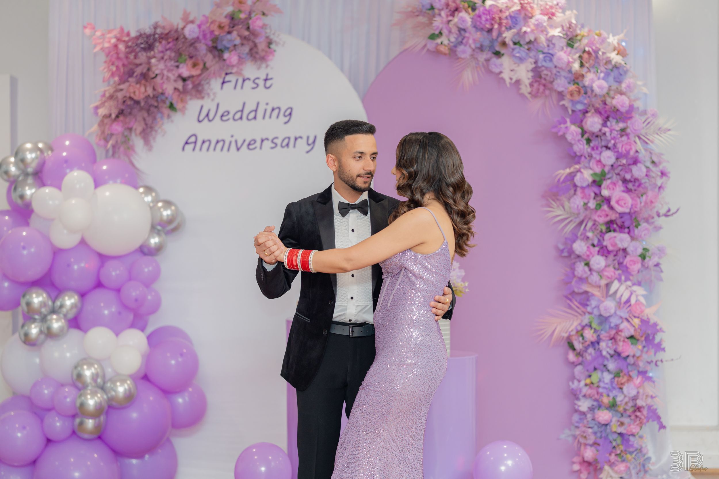 Wedding couple poses under purple floral arch with balloons and romantic lighting at their first wedding anniversary celebration.