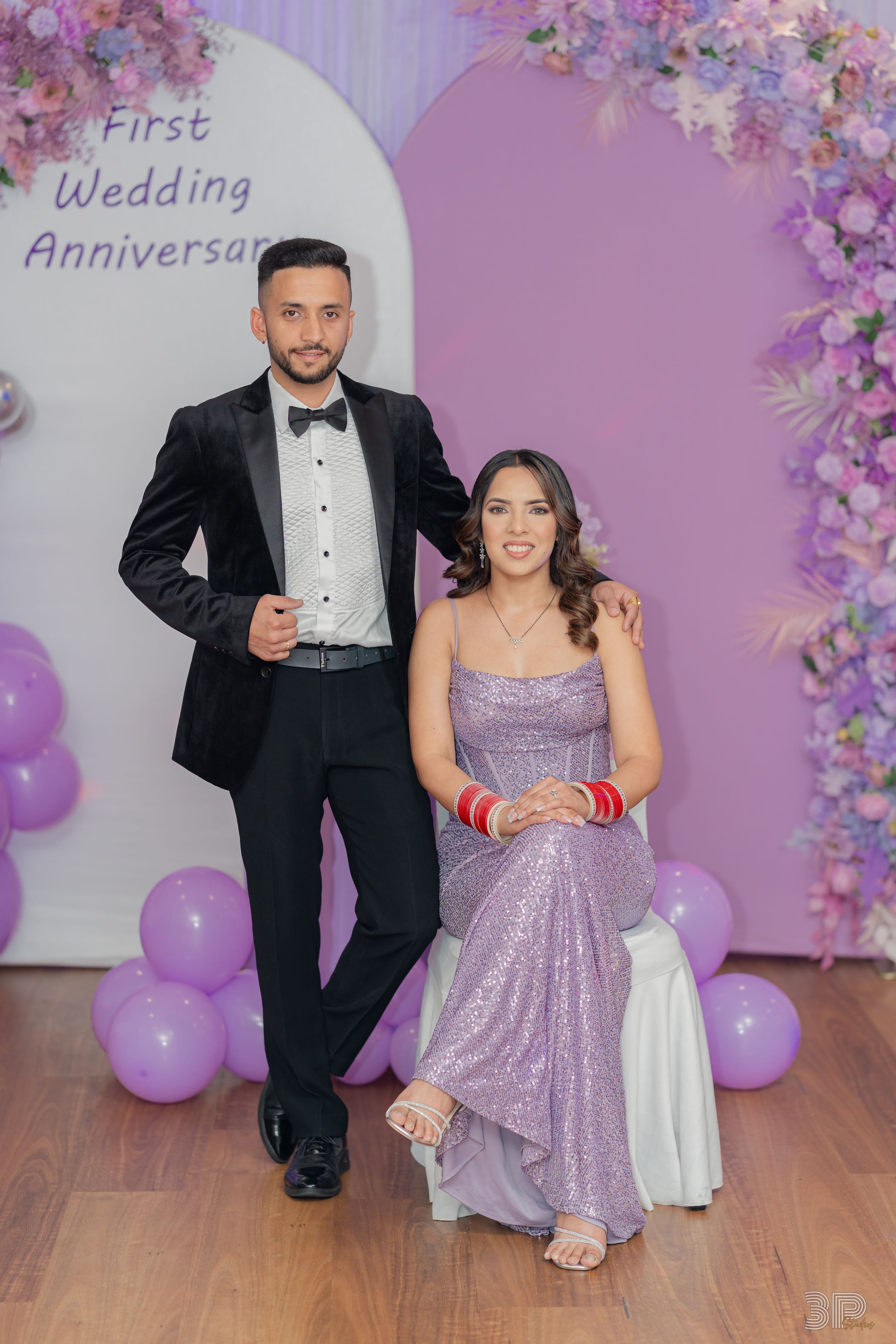 A couple poses in formal attire against a purple floral backdrop with balloons at their wedding anniversary celebration.