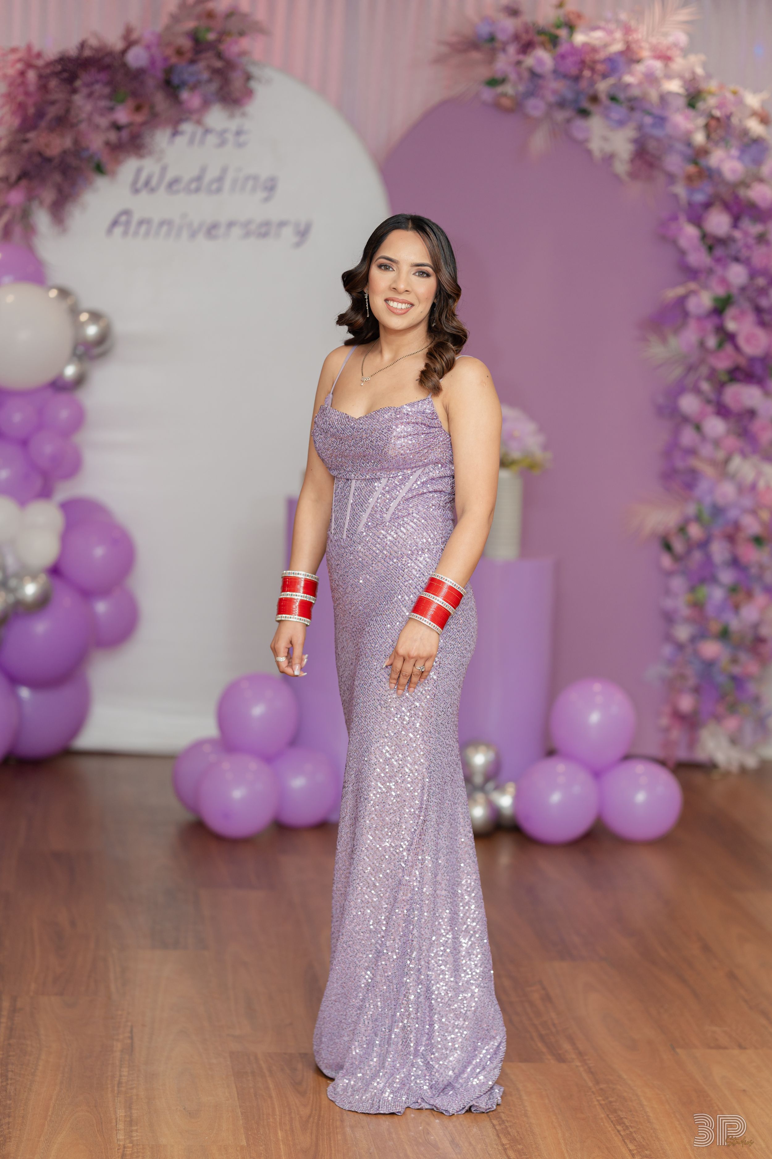 A person in a sparkly lavender gown poses in front of a purple balloon arch and floral wedding anniversary backdrop.