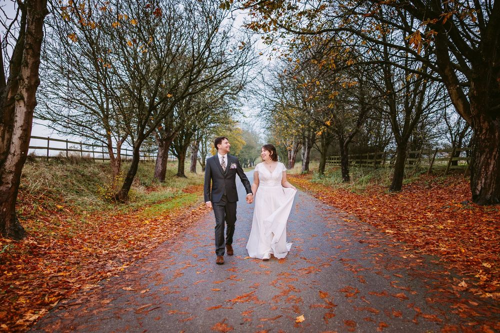 A couple in wedding attire embraces in a scenic field at sunset with dramatic cloudy skies.