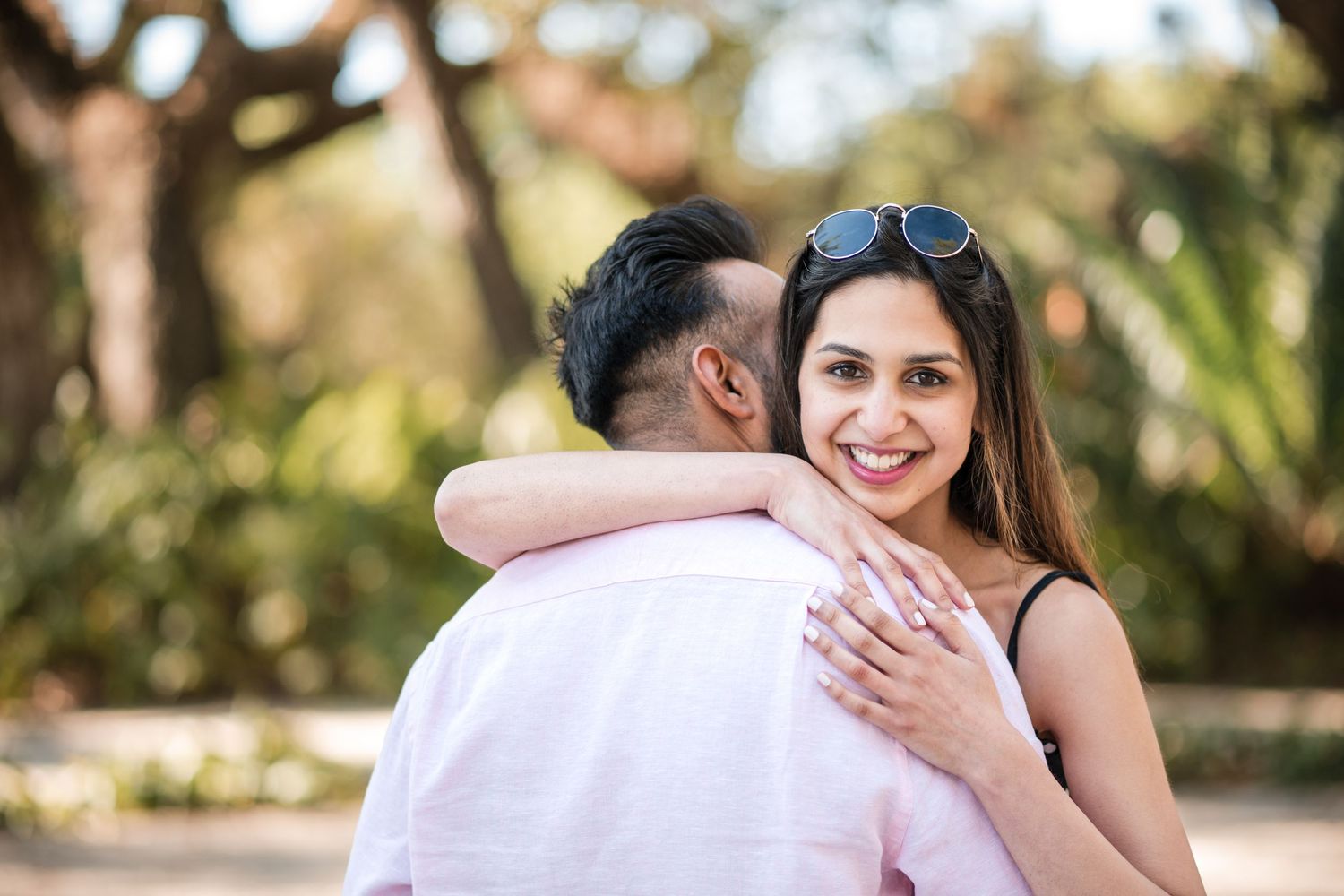 A smiling woman with sunglasses hugs a man outdoors in a park setting, showcasing a ring on her hand.