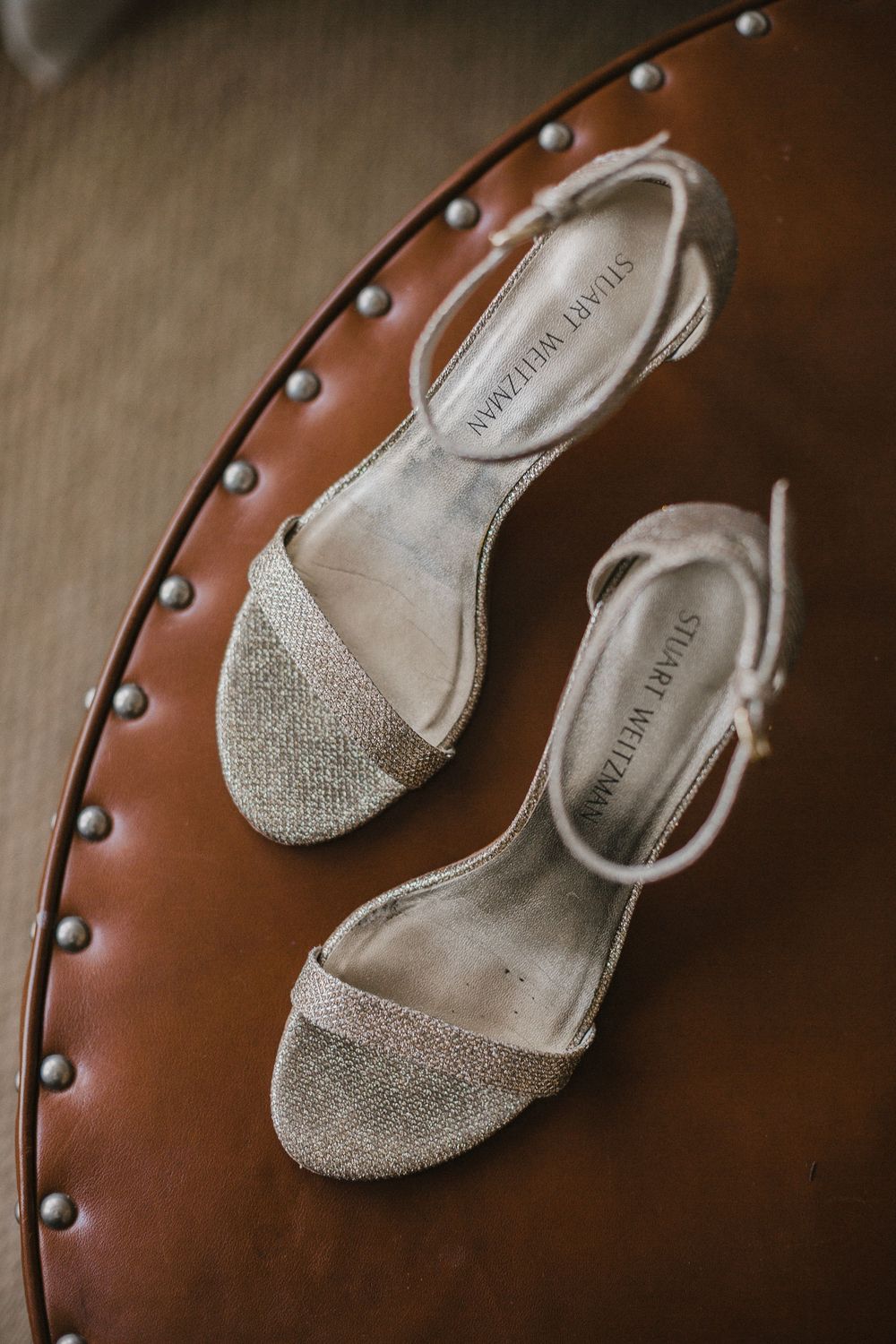 Sparkly silver wedding sandals displayed on a brown leather chair with decorative studs.