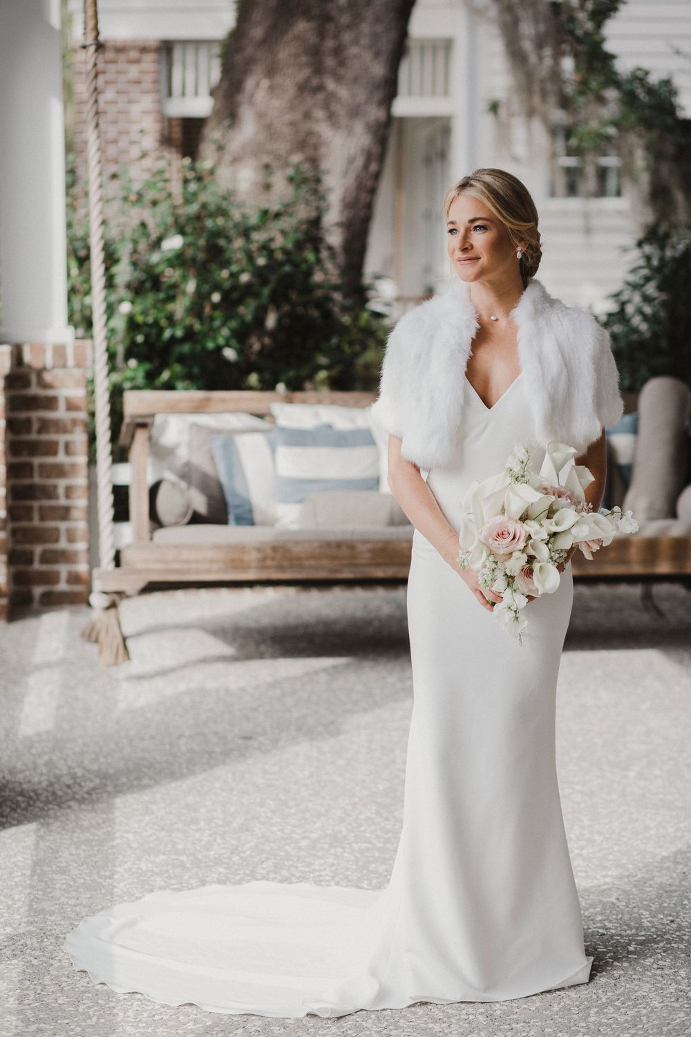 Elegant bride in white gown with fur shawl holding bouquet on a porch.