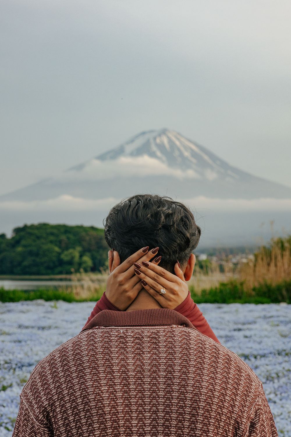 Person in knit sweater views Mount Fuji from a scenic overlook with blooming flowers in the foreground.