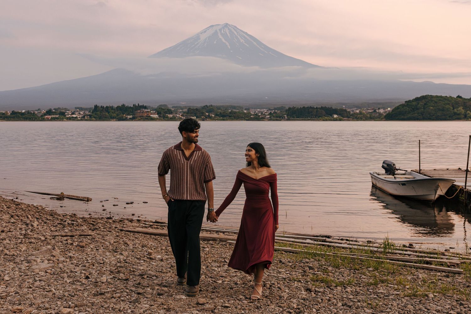 A couple walks along the shore of Lake Kawaguchiko with Mount Fuji rising majestically in the background at sunset.