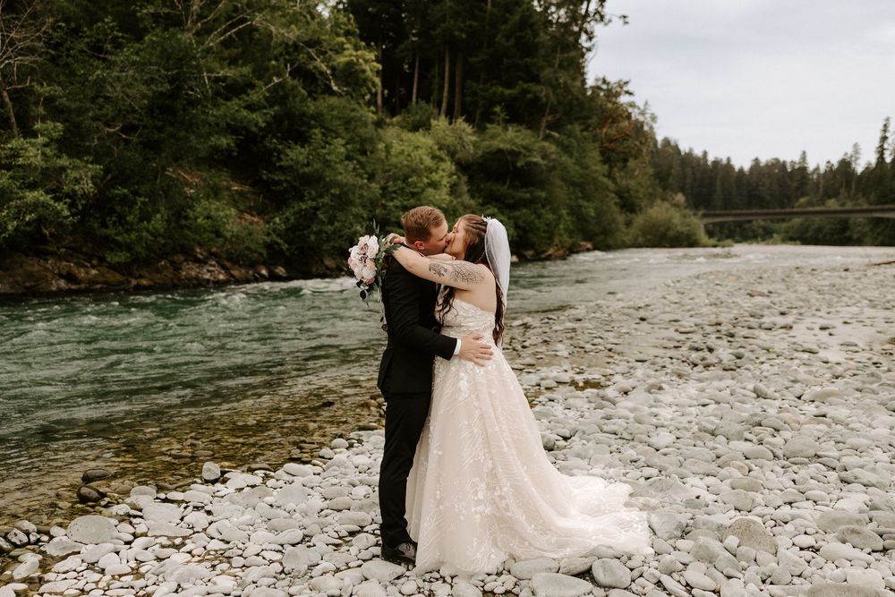 Romantic wedding photos captured on a scenic rocky beach surrounded by evergreen forest and mountains.