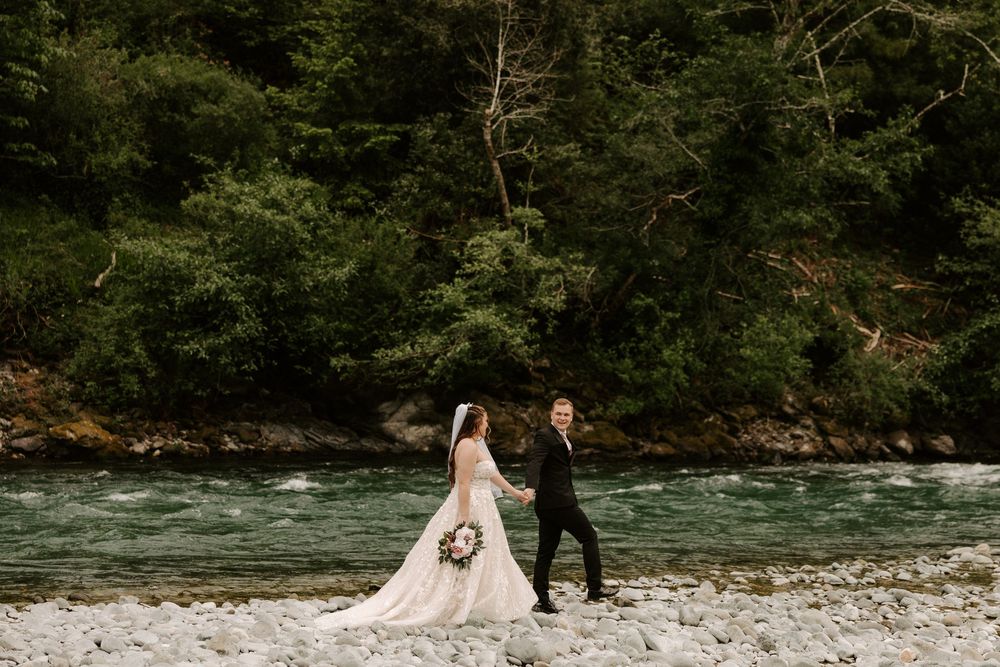 A wedding couple walks along a scenic riverbank surrounded by lush forest and flowing water.