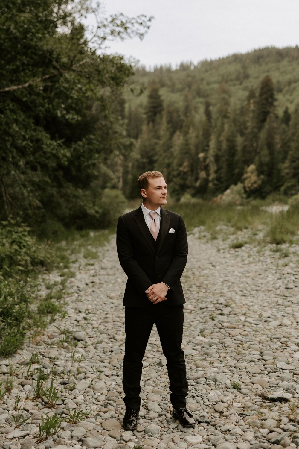 A person in formal attire stands on rocky terrain surrounded by mountain scenery.