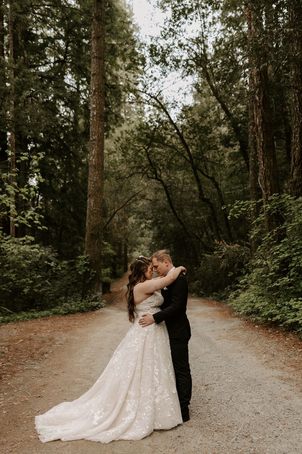 A romantic moment captured on a tree-lined forest path with soft moody lighting and natural surroundings.