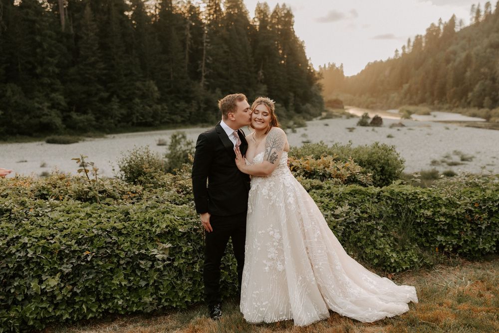 A couple in wedding attire embraces during golden hour in a scenic mountain landscape with evergreen trees.