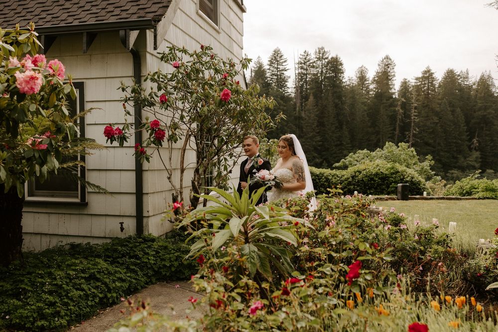 A romantic garden scene with climbing roses and lush greenery surrounds a white house on a summer day.