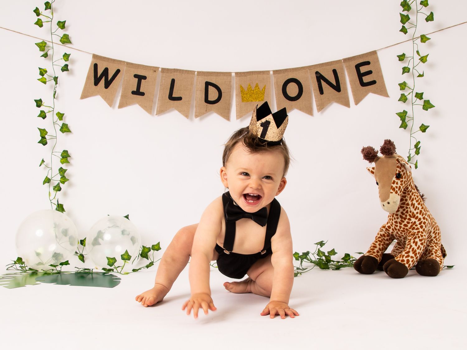 A baby crawling on a white backdrop with green vine decorations and a rustic banner hanging above.