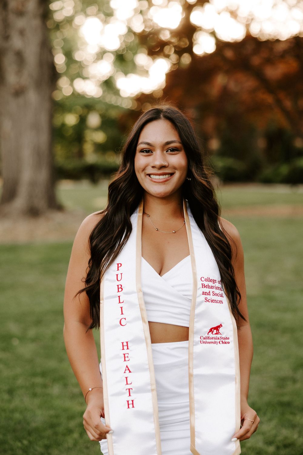 Graduate in white dress and ceremonial stole smiles while posing outdoors during graduation photo session.