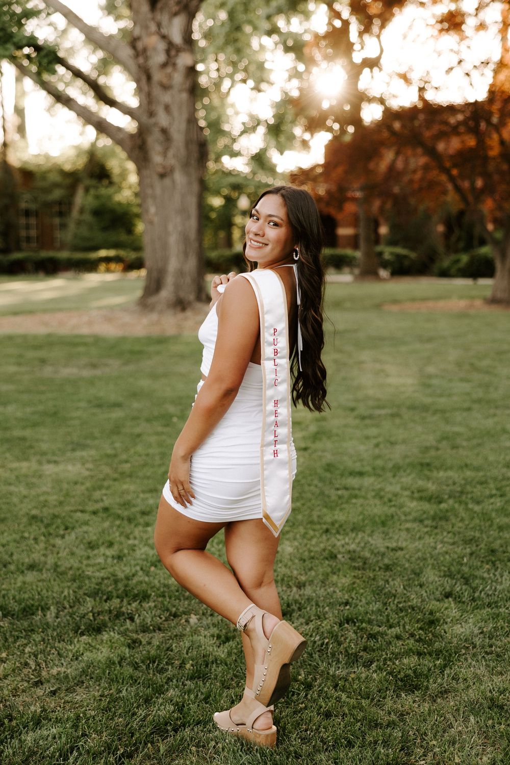 A person in a white dress poses playfully on a grassy hill during golden hour sunset.