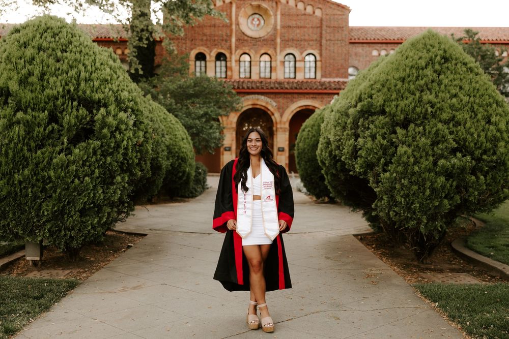 A person poses in front of a brick building surrounded by lush green bushes and landscaping.