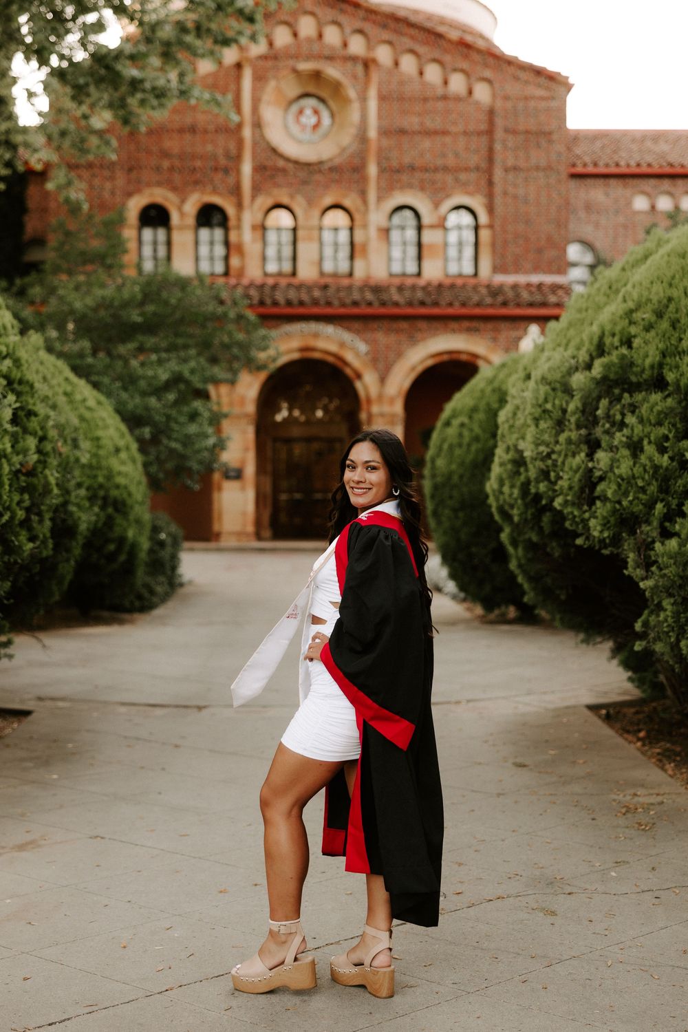 A graduate poses in front of a red brick university building with arched windows and architectural details.