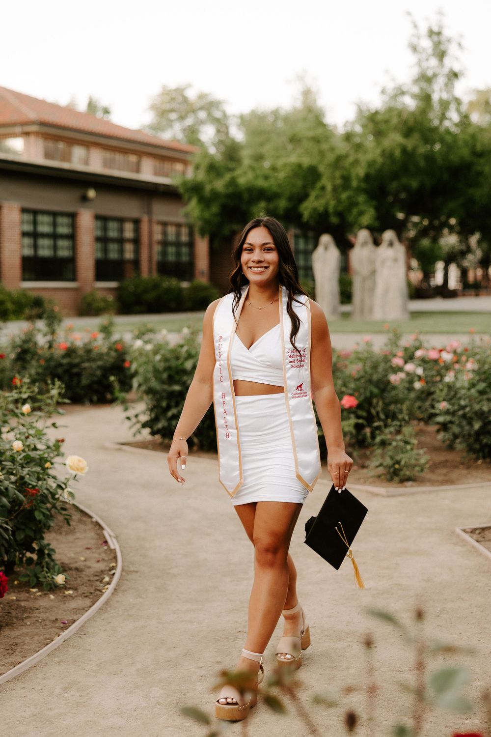 Woman in white dress walking through a garden path with brick buildings and rose bushes in the background.