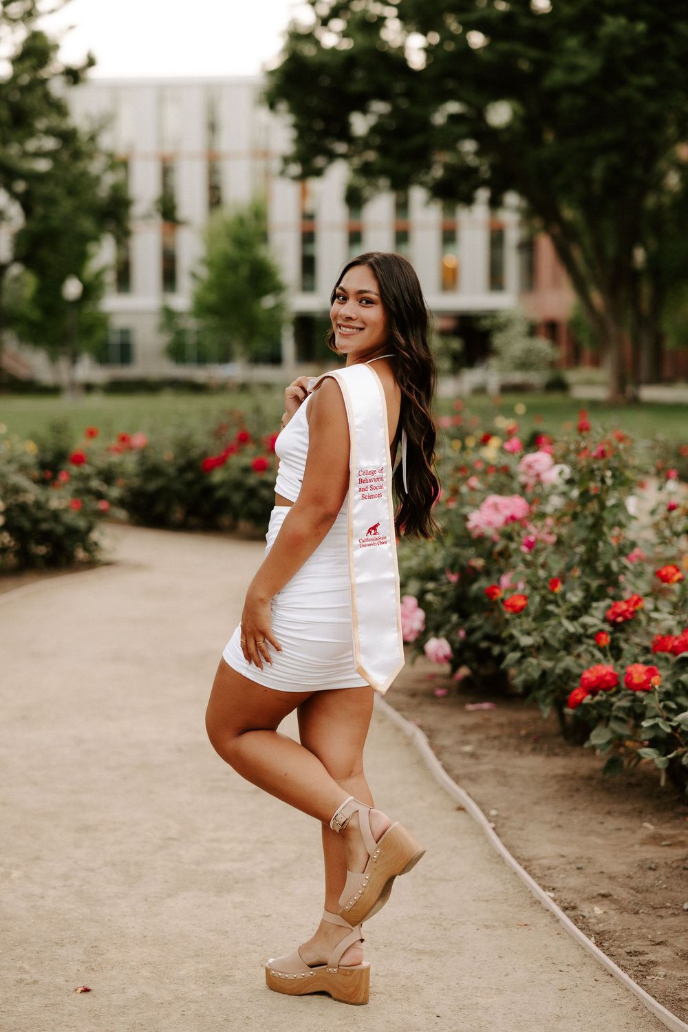 A person poses in a white tank top and shorts near blooming red rose bushes on a garden path.