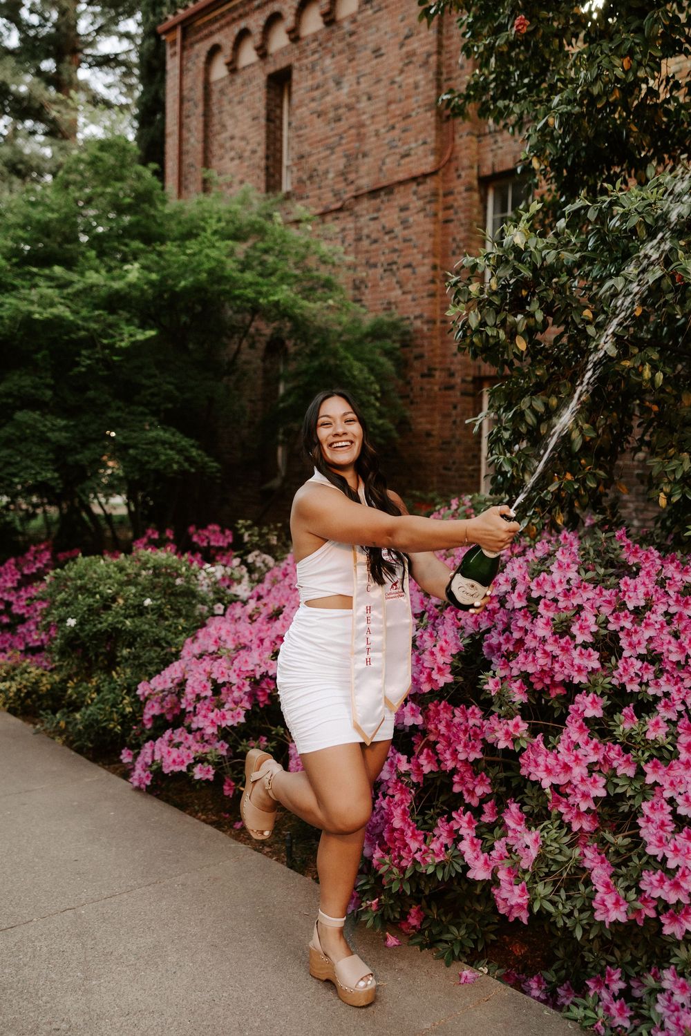 Person in white outfit poses playfully near vibrant pink azalea bushes against brick building backdrop.