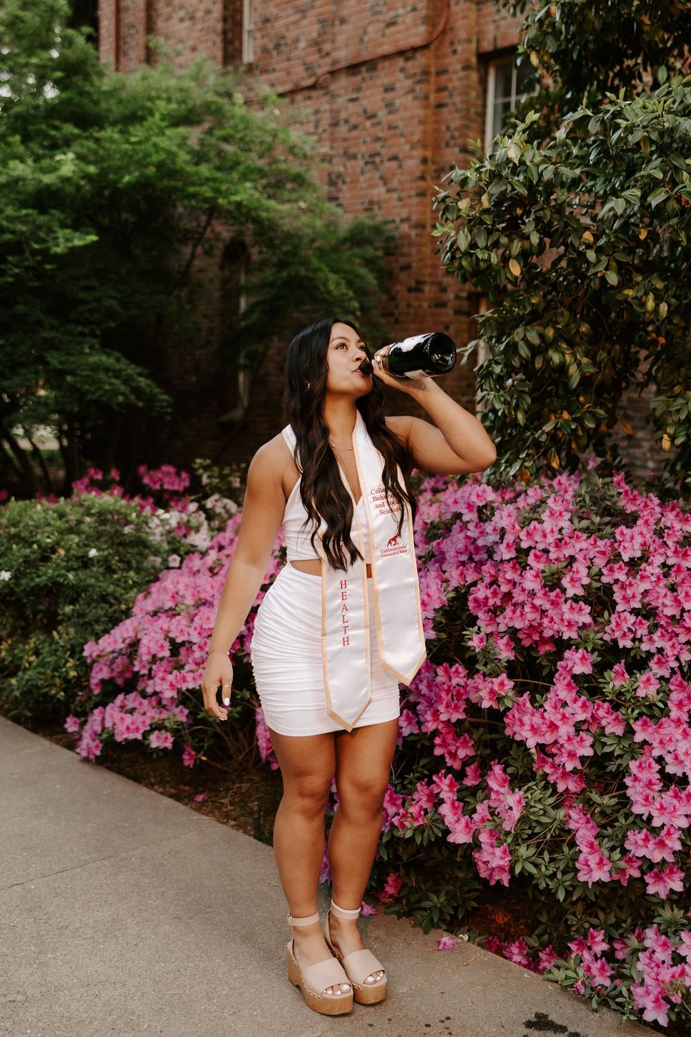 A person in a white top poses near vibrant pink azalea bushes against a brick wall backdrop.