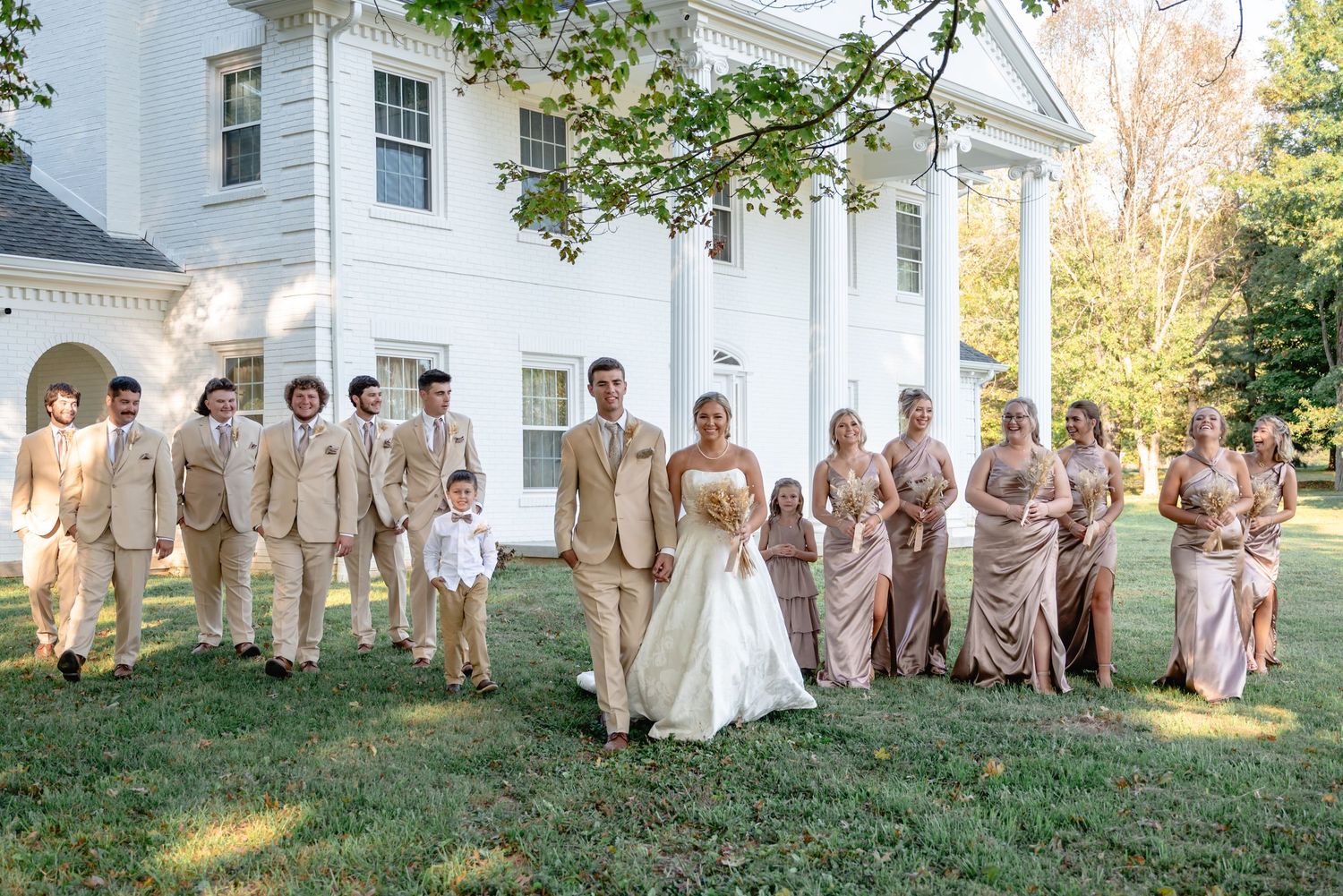 Wedding party in gold and blush gowns poses on a lawn before a white colonial mansion.