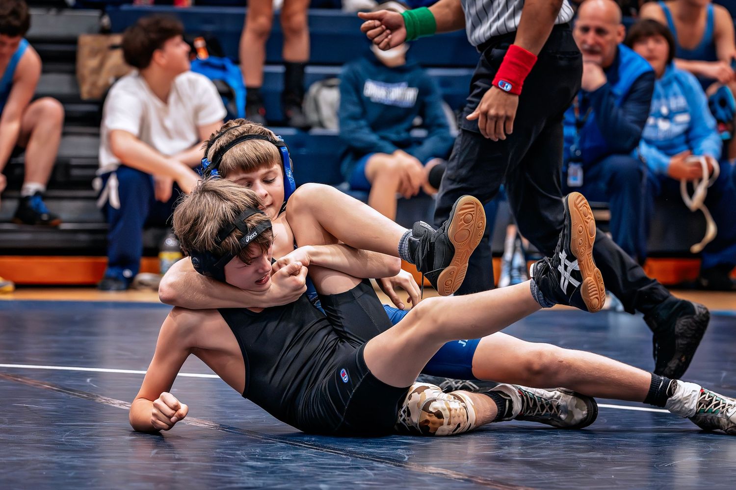 High school wrestlers compete in a tournament match on blue wrestling mats with spectators watching in the background.