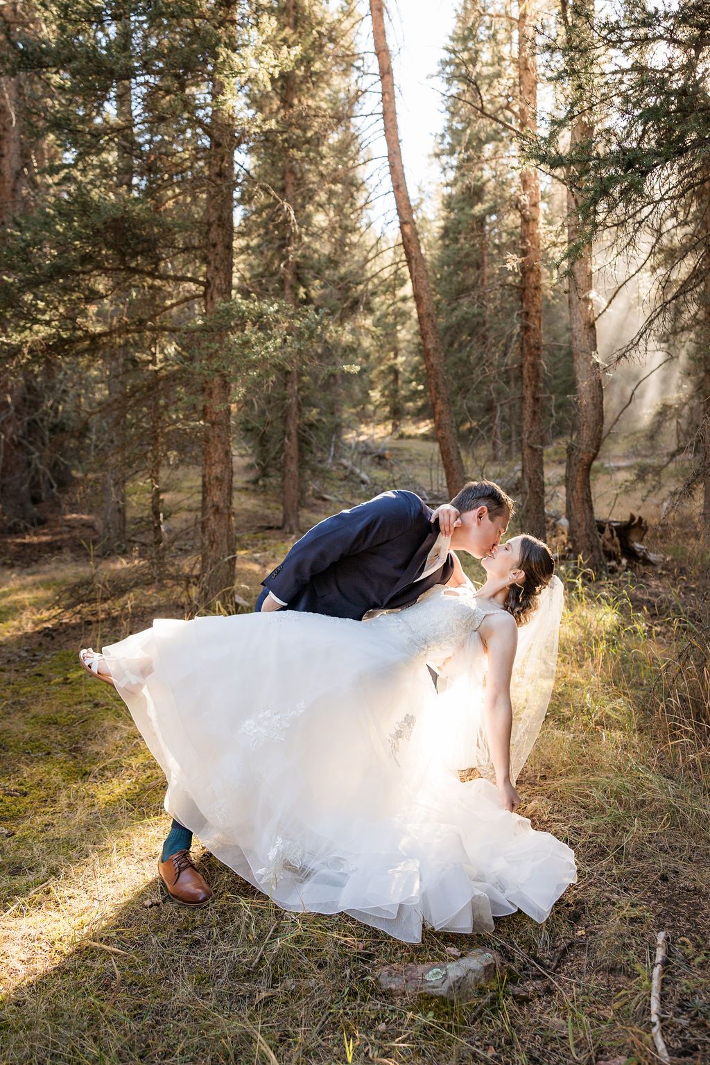 A couple in wedding attire shares romantic moments in a sunlit forest setting with tall pine trees.