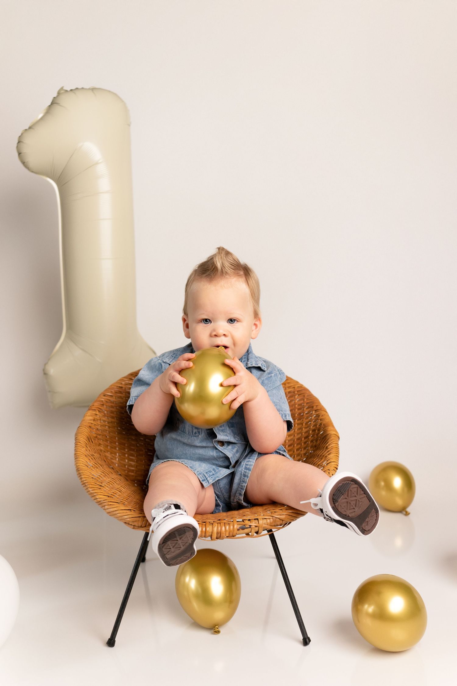 A baby in denim overalls sits on a woven chair with golden balloons in a minimalist photo series.
