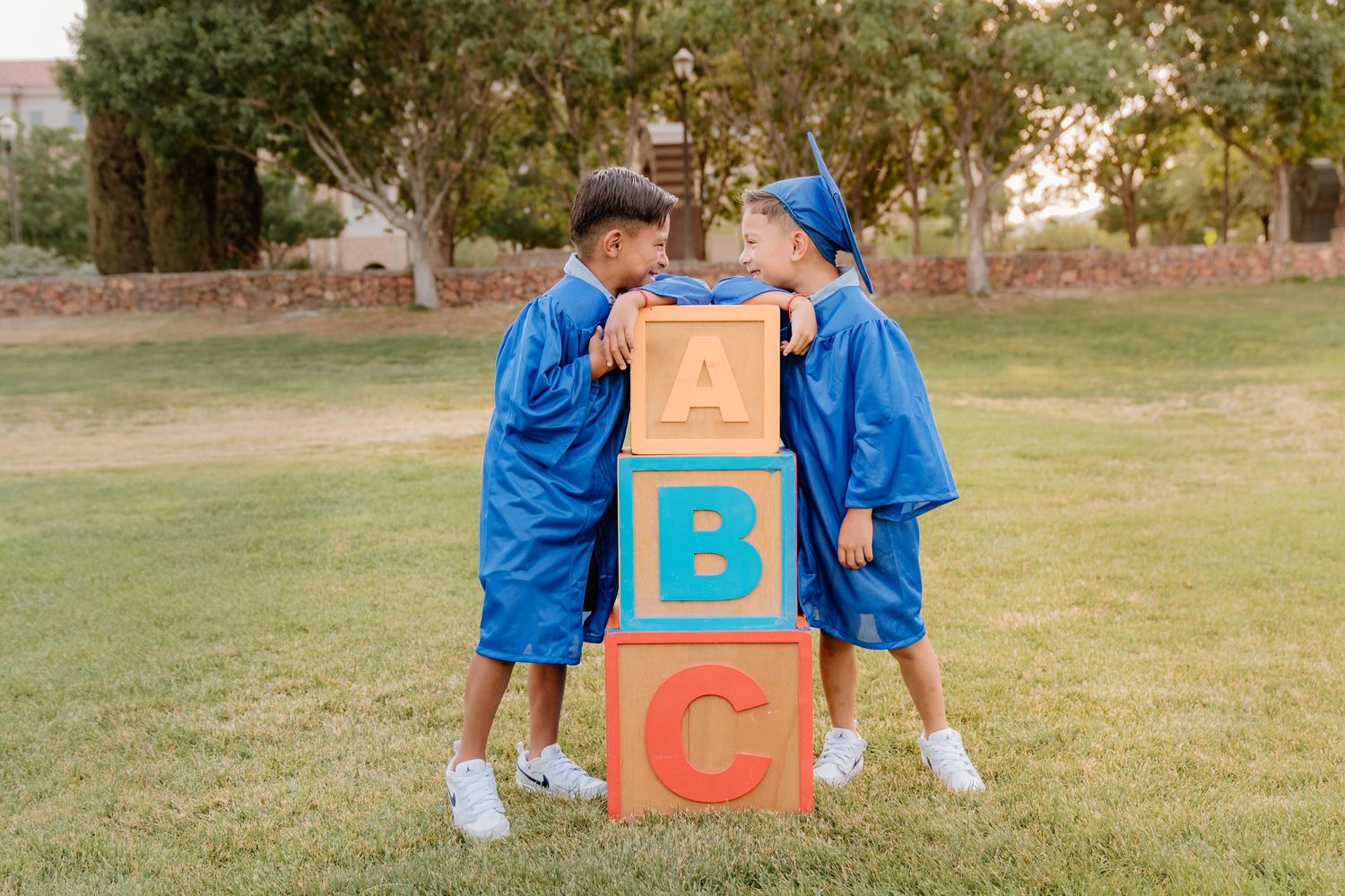 Two boys smile and laugh together in a grassy field, wearing kindergarten graduation gowns.