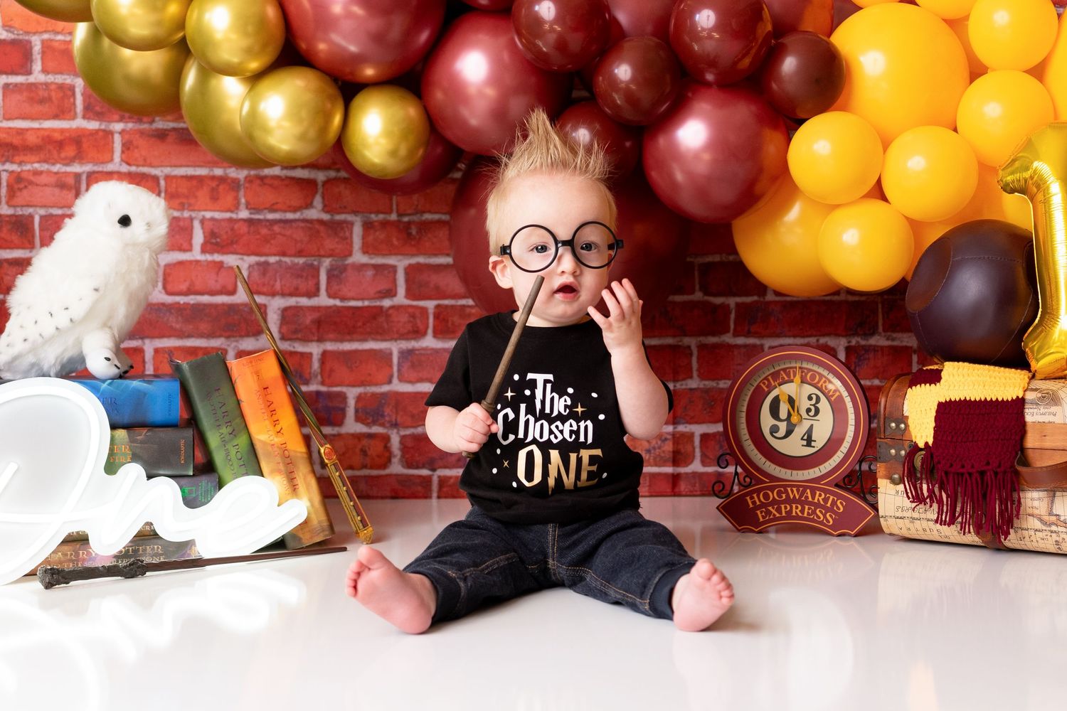 Baby sits in blue basin with colorful balloon arch, vintage suitcases, and rocking horse against brick wall backdrop.