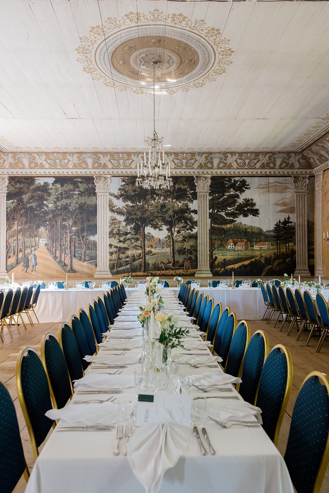 Long banquet tables with blue chairs set for formal dining in an elegant room with ornate ceiling and wall murals.