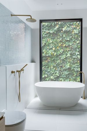 Minimalist bathroom featuring freestanding tub, gold fixtures, and large window with green foliage view.