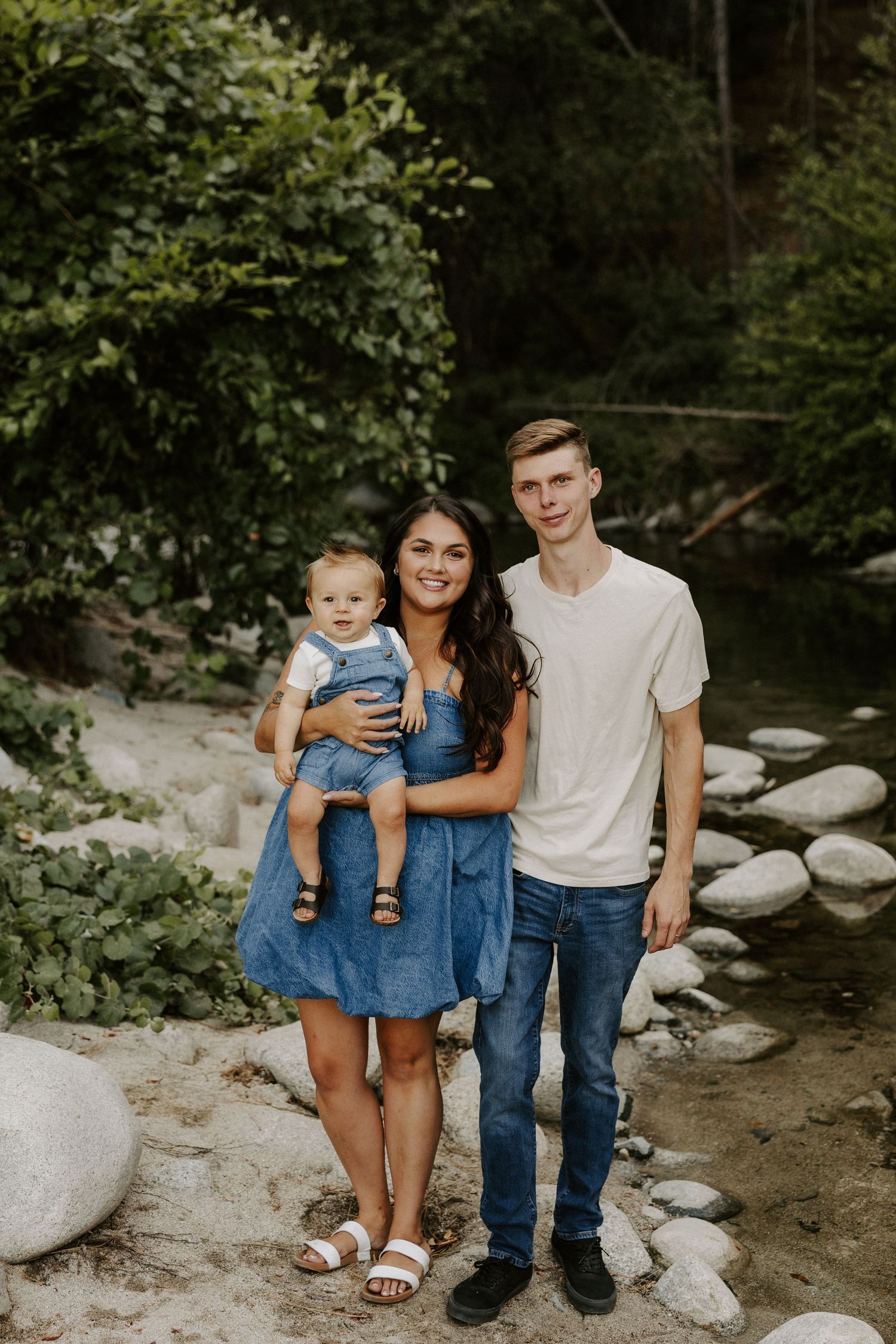 Family in coordinating denim outfits poses together on rocky riverbank in natural outdoor setting.