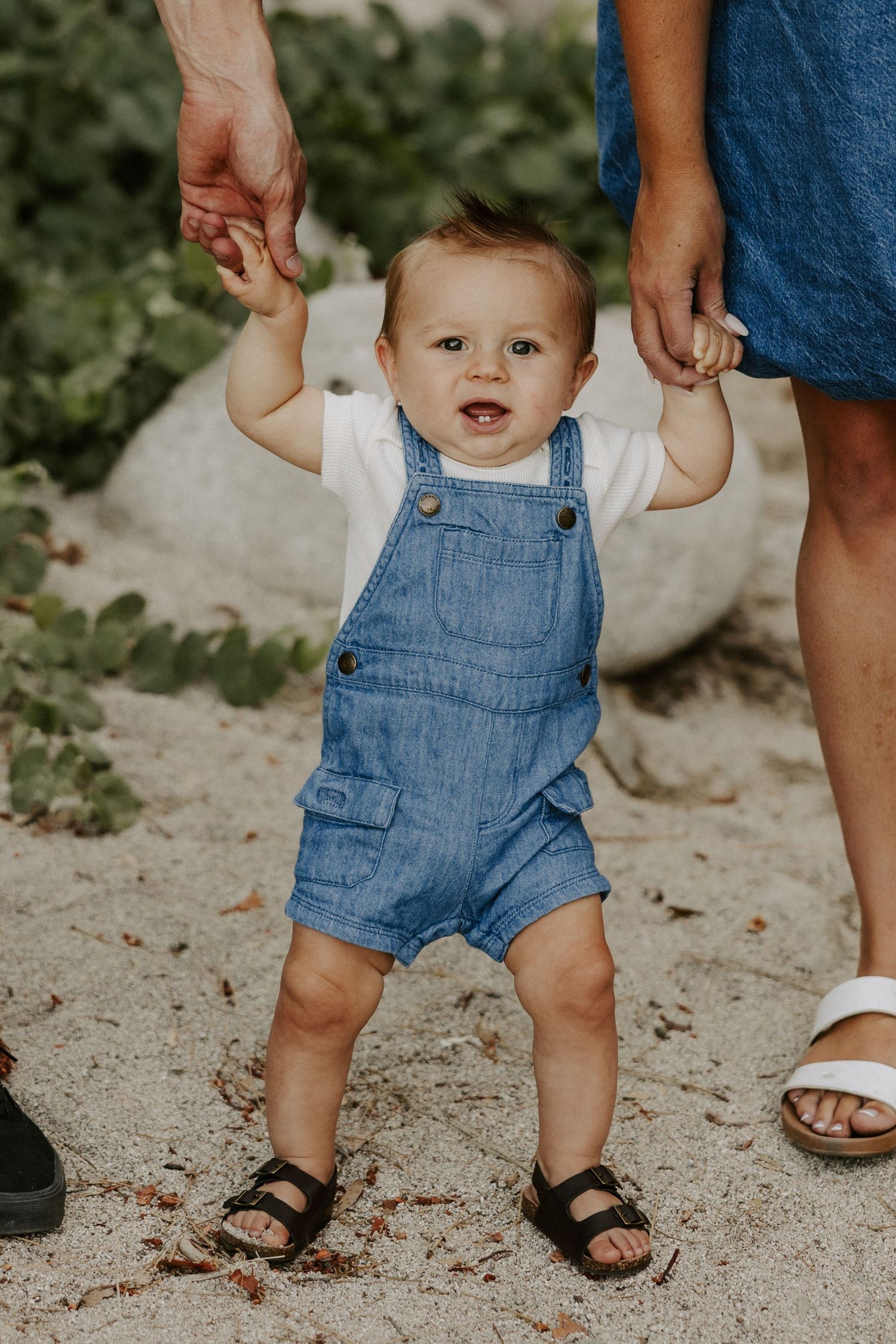 Small child in denim overalls takes first steps while holding hands with adult on sandy ground.