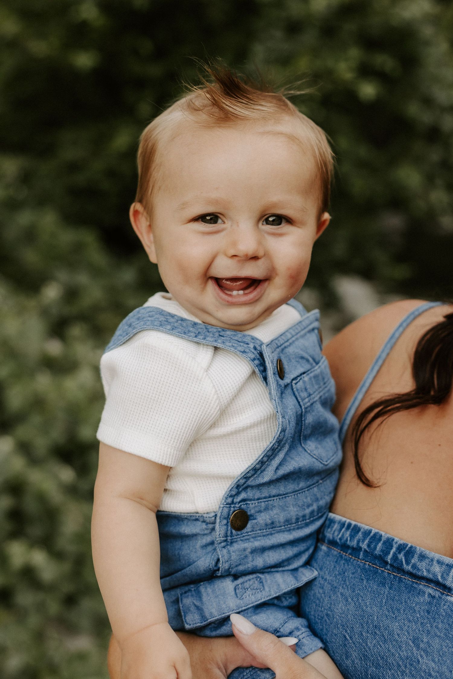 A baby in denim overalls and white shirt smiles while being held outdoors with greenery in the background.
