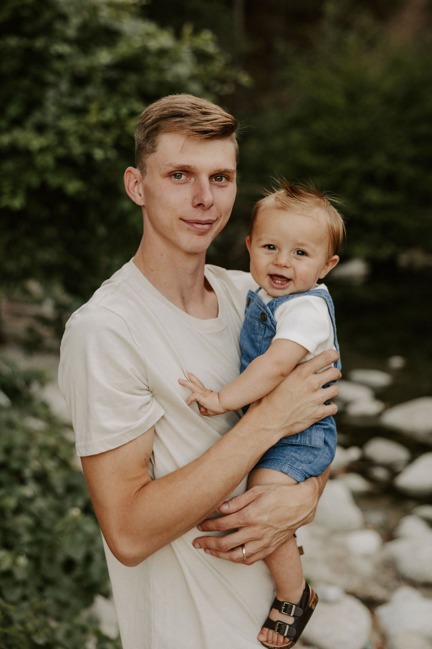 A toddler is held and embraced while wearing a white shirt in an outdoor setting with greenery.