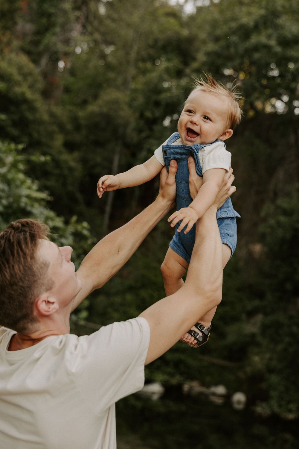 A joyful parent lifts and plays with a smiling baby wearing blue against a green forest backdrop.