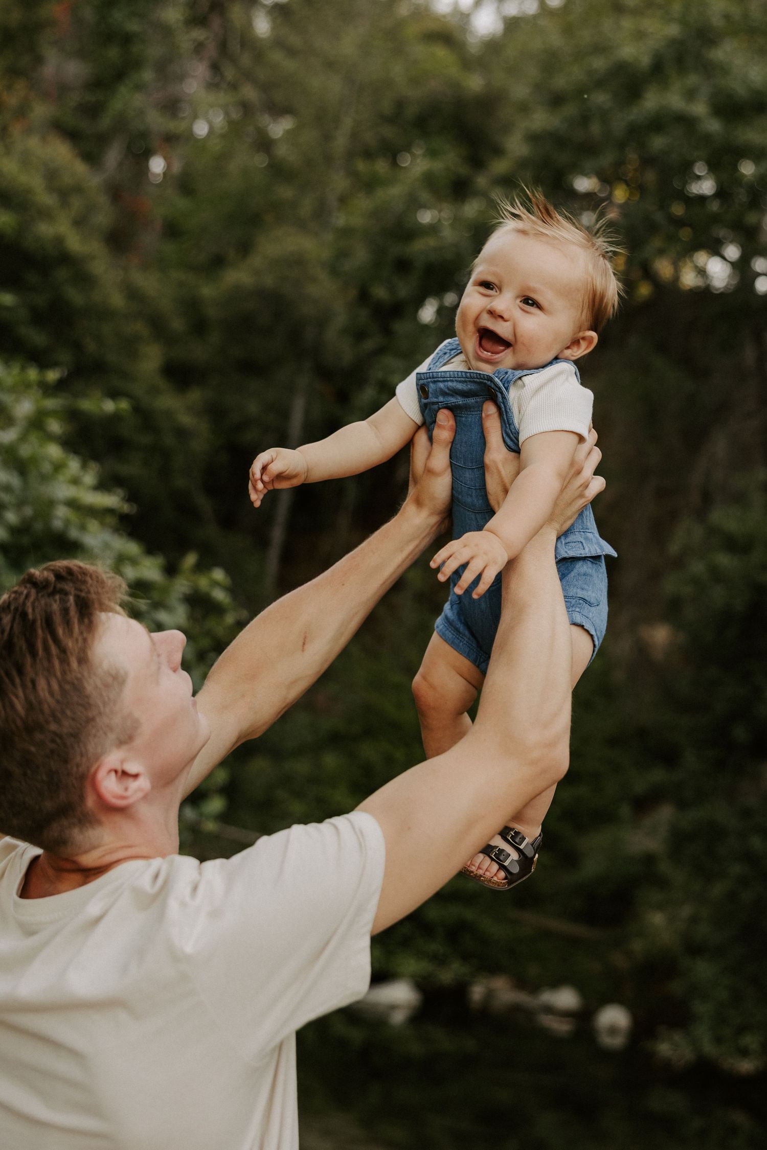 A joyful parent lifts and plays with a smiling baby wearing blue against a green forest backdrop.