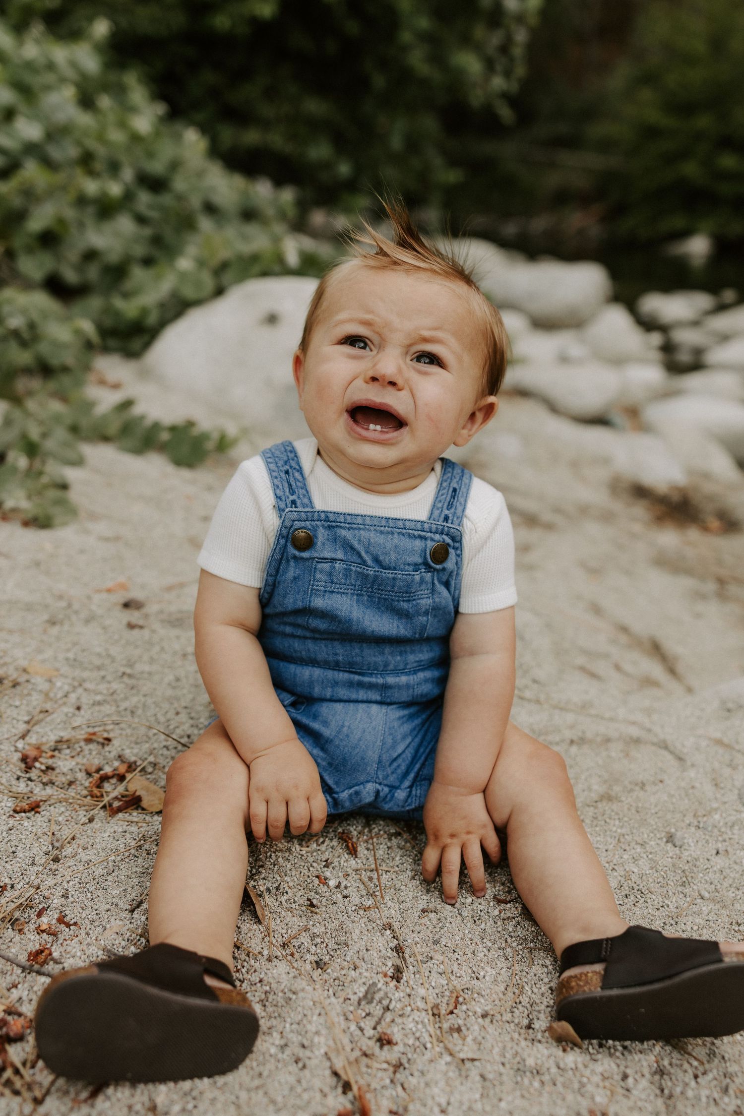 A baby wearing denim overalls and a white shirt sits on a sandy beach, smiling and playing in three progressive shots.