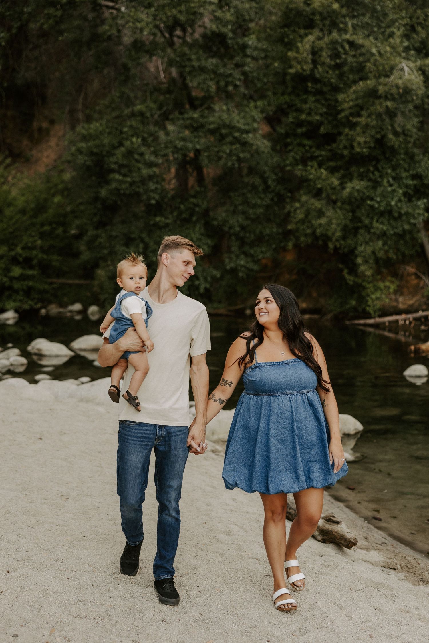 A family walks together along a sandy riverbank surrounded by green trees and rocks.