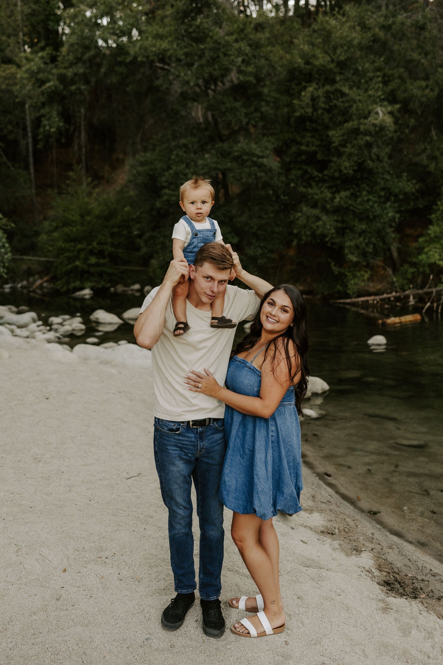 Family shares candid moments and laughter while sitting together on sandy riverbank surrounded by trees.