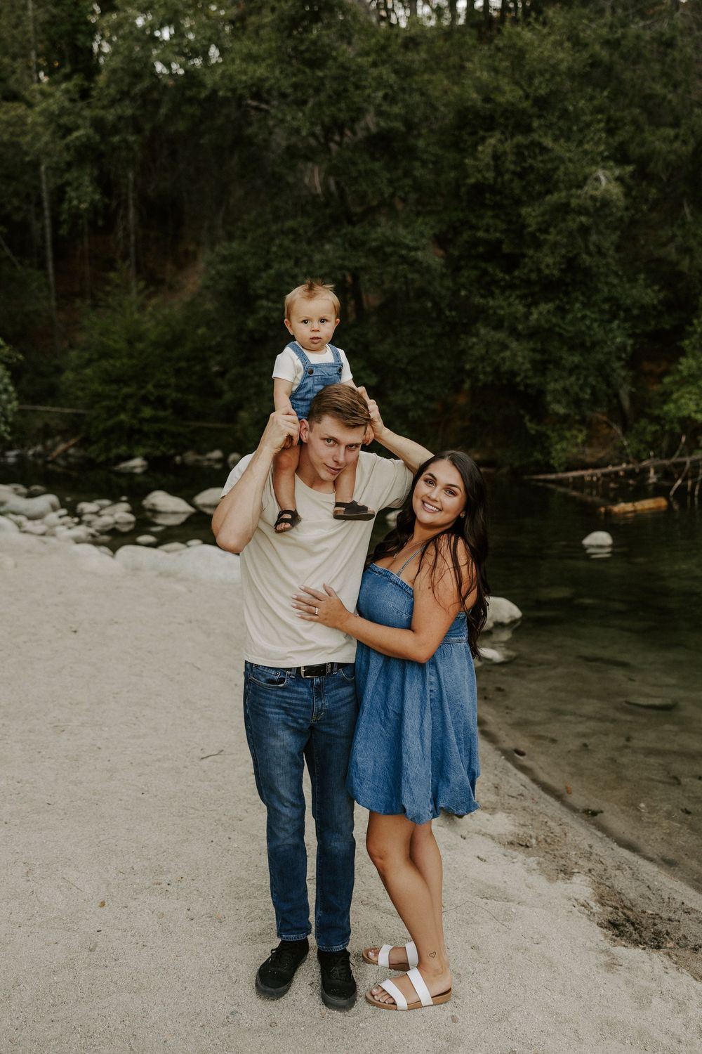 Family shares candid moments and laughter while sitting together on sandy riverbank surrounded by trees.