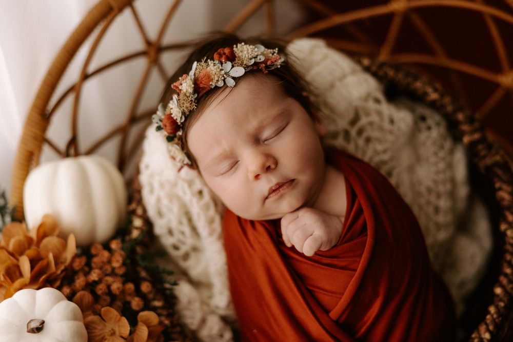 Sleeping baby in rust-colored wrap nestled in wicker basket with white cushion and autumn pumpkin decorations.